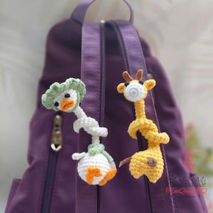 Peut inclure: Deux breloques faites à la main en crochet représentant des animaux : un canard avec un chapeau vert et une girafe. Le canard est blanc avec des touches d'orange, et la girafe est jaune avec des taches marron. Elles sont attachées à un sac à dos violet.