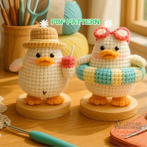 Può includere: Due figurine di anatra all'uncinetto. Un'anatra indossa un cappello di paglia e tiene in mano una bevanda rosa. L'altra anatra indossa occhiali da sole rosa e un salvagente a righe blu e gialle. Entrambe le anatre hanno becchi e zampe arancioni. Il testo "PDF PATTERN" è visibile.