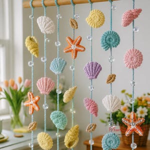 May include: A wall hanging featuring multiple vertical light blue cords suspended from a wooden rod. The cords are decorated with various crocheted sea creatures including starfish, conch shells, and scallops in pastel colors, interspersed with clear beads.