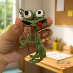 Può includere: Un portachiavi a forma di rana all'uncinetto verde fatto a mano con una lingua rossa e occhi bianchi e neri. La rana ha un design stravagante ed è attaccata a una clip in metallo color argento. Lo sfondo è sfocato.