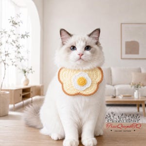 May include: A white cat wearing a crocheted pet bib featuring a central fried egg design with a yellow yolk and white egg white, surrounded by a scalloped light yellow border with a thin brown edge. The text TrucQuynhTQ CROCHET PATTERN is visible.