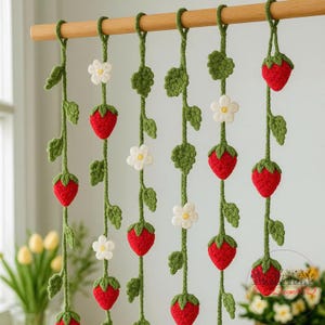 May include: Crocheted strawberry garland with green vines, leaves, and white flowers. The red strawberries are evenly spaced along the vines, creating a charming, handmade decorative piece. The garland hangs from a wooden rod.