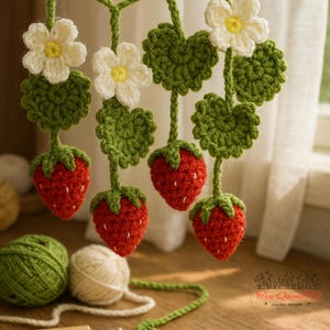 May include: Crocheted strawberry garland featuring red strawberries, green leaves, and white flowers with yellow centers. The handmade garland is suspended, with yarn balls in the foreground. The overall theme is a spring or summer decoration.