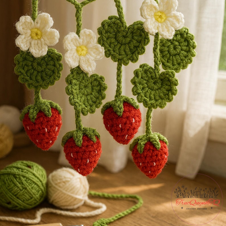 May include: Crocheted strawberry garland featuring red strawberries, green leaves, and white flowers with yellow centers. The handmade garland is suspended, with yarn balls in the foreground. The overall theme is a spring or summer decoration.