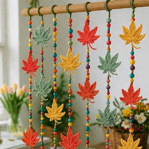 May include: A decorative hanging featuring crocheted maple leaves in shades of red, green, and yellow. The leaves are strung on beaded strands with colorful wooden beads. The leaves are attached to a wooden rod with green twine.