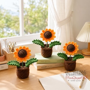 May include: Three crocheted sunflowers in brown pots with green leaves. The sunflowers have orange petals and brown centers. The pots are sitting on a desk with books and a notepad.