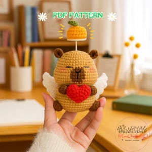May include: A crocheted bear-like figure with a red heart, white wings, and a small orange pumpkin on its head. The figure is tan with brown accents and closed eyes. The text "PDF PATTERN" is visible.