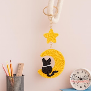 May include: A handmade crochet keychain featuring a yellow star and a yellow crescent moon with a black cat silhouette. The keychain is attached to a gold-colored ring. The background includes a clock, pencils, and a ruler.