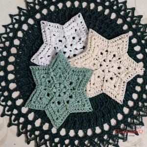 May include: Three crocheted star-shaped coasters in white, cream, and green, arranged on a dark green, lacy doily. The stars have a floral design in the center. The doily has a scalloped edge.