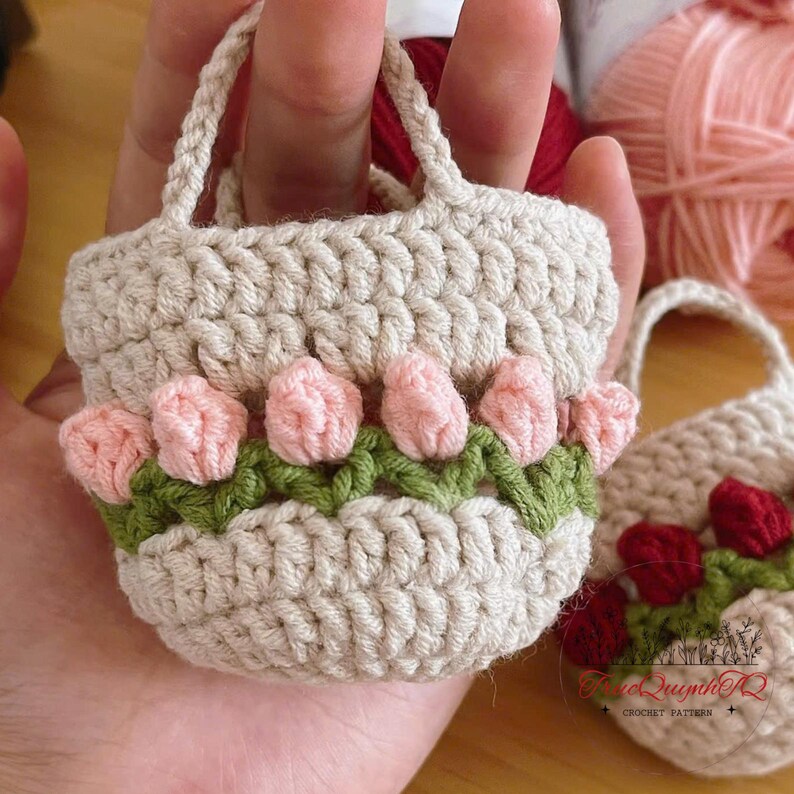 May include: A small, crocheted beige handbag with pink flower accents and green leaves. The miniature bag has a handle and is decorated with several pink flowers and green leaves. The bag is handmade and has a textured appearance.