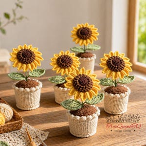 Puede incluir: Una colección de girasoles de ganchillo hechos a mano en macetas pequeñas. Los girasoles tienen pétalos amarillos y centros marrones, con hojas y tallos verdes. Las macetas son beige con un borde festoneado. El arreglo está sobre una superficie de madera.