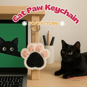 Può includere: Un portachiavi a forma di zampa di gatto all'uncinetto con centro nero e cuscinetti rosa e bianchi è appeso a una superficie di legno. Il testo "Cat Paw Keychain" e "PDF PATTERN" sono visualizzati sopra. Un gatto nero è sullo sfondo.