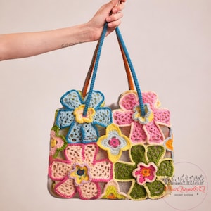 May include: A colorful crochet tote bag featuring large floral designs in shades of blue, pink, yellow, and green. The bag has blue and orange rope handles. The bag is held up by a hand.
