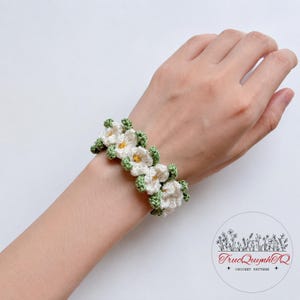 May include: A white and green crochet bracelet featuring a floral design. The bracelet is made of white flowers with yellow centers and green leaves. The bracelet is worn on a wrist.