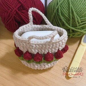 May include: A crocheted basket with a handle, decorated with red strawberry-like embellishments and green leaves. The basket is filled with a white bar of soap. Yarn in red and green are in the background.