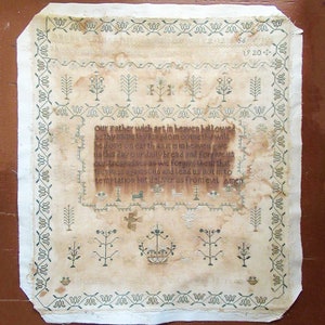 以下が含まれることがあります： 宗教的なテキストが刺繍されたアンティークのサンプラー。装飾的なボーダーと様々なステッチモチーフが特徴です。「主の祈り」と書かれたテキストは、緑と茶色の糸で、色あせた、古びた外観です。