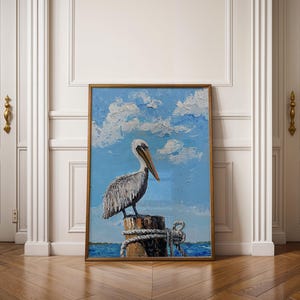 Pode incluir: Pintura emoldurada de um pelicano empoleirado em uma estaca de madeira. O pelicano tem penas brancas e cinzentas, um bico longo, em um fundo de céu azul com nuvens brancas. A pintura está em uma moldura dourada.