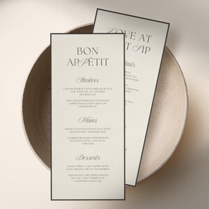 Może przedstawiać: Dwa eleganckie menu z czarnymi obramowaniami, z frazami "Bon App&eacute;tit" i "Love at First Sip." Menu zawiera przystawki, dania gł&oacute;wne i desery. Karty są umieszczone w beżowej misce, tworząc wyrafinowaną prezentację.