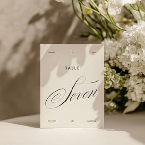 以下が含まれることがあります： オフホワイトの長方形のテーブルナンバーカード。TABLE SEVENの文字がサンセリフ体とエレガントな筆記体で印刷されています。カードにはAUGUST 22 2028の日付と、WHITNEY AND SEBASTIANの名前が小さな黒い文字で記載されています。