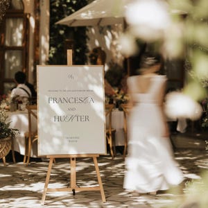 Może przedstawiać: Drewniany sztalug prezentuje biały znak z eleganckim czarnym tekstem. Znak głosi "Francesca and Hunter" z datą "10.20.25" poniżej. Scena rozgrywa się na zewnątrz, z rozmytą postacią w białej sukni w tle.