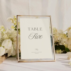 Może przedstawiać: Złota ramka z numerem stołu z napisem "TABLE Five" w eleganckiej czcionce. Znak zawiera r&oacute;wnież imiona "Francesca & Hunter" i datę "10.20.25". Białe kwiaty zdobią tło.