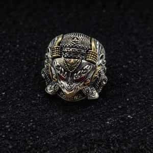 Peut inclure: Une bague en métal argenté représentant un visage de créature stylisé et détaillé, avec des accents dorés sur les cornes et le front. La créature possède des yeux rouge foncé et des motifs gravés complexes sur toute la surface de l'anneau.