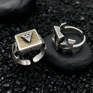 Puede incluir: Dos anillos de plata con una cara cuadrada con un triángulo dorado con un diseño de ojo. Los anillos tienen una banda ajustable y están colocados sobre un fondo oscuro y texturizado. Los anillos tienen un símbolo ankh en el lateral.