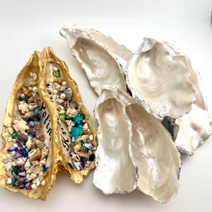 May include: A collection of oyster shells, some natural and some painted gold. One gold shell is filled with small seashells, beads, and turquoise turtle figurines. The word "Polina" is written on the gold shell. The other shells are white.