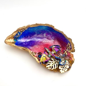 May include: A decorative oyster shell with a gold-painted rim and a colorful interior. The interior features a blend of blue, purple, and pink hues, with iridescent dragon figurines and gold leaf accents. Small gemstones and white leaf-shaped embellishments add detail.