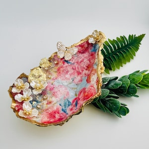 May include: An oyster shell trinket dish with a floral design in pink, red, and blue. The shell is adorned with gold accents, including a butterfly and flower embellishments. A decorative piece for jewelry or small items.