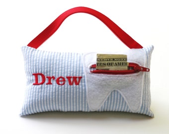 May include: A rectangular light blue and white striped pillow with a red fabric handle. The name Drew is embroidered in red on the front. A white felt tooth shape is appliqued on the right side, featuring a red zippered pocket containing a dollar bill.