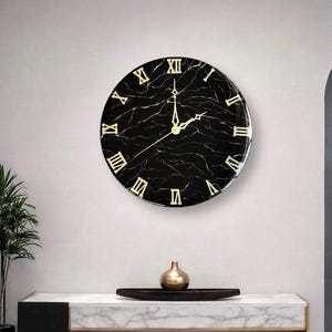 May include: A round wall clock with a black marble-like face and gold Roman numerals. The clock hands are gold. The clock is mounted on a light gray wall. The clock is a decorative item.
