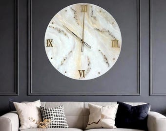 May include: A large, round wall clock with a cream-colored face and gold Roman numerals. The clock hands are gold, and the face has a marbled effect with embedded decorative elements. The clock is mounted on a dark gray wall.