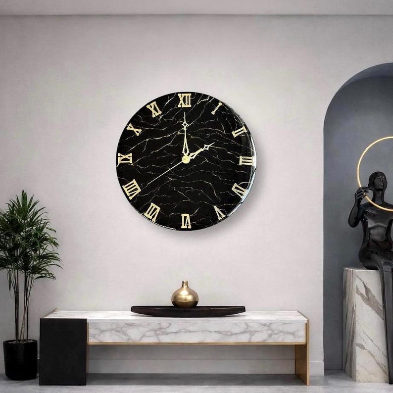 May include: A round wall clock with a black marble-like face and gold Roman numerals. The clock hands are gold. The clock is mounted on a light gray wall. The clock is a decorative item.
