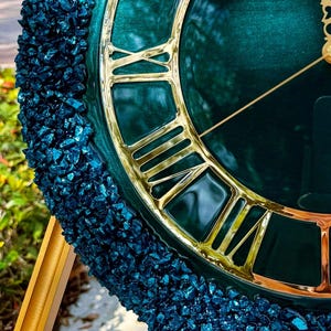 K&ouml;nnte beinhalten: Eine runde Wanduhr mit einem tief smaragdgr&uuml;nen Zifferblatt, polierten goldenen r&ouml;mischen Ziffern und passenden d&uuml;nnen goldenen Zeigern. Der &auml;u&szlig;ere Rand ist mit einer texturierten Schicht aus dunkelblauen Glassplittern besetzt.