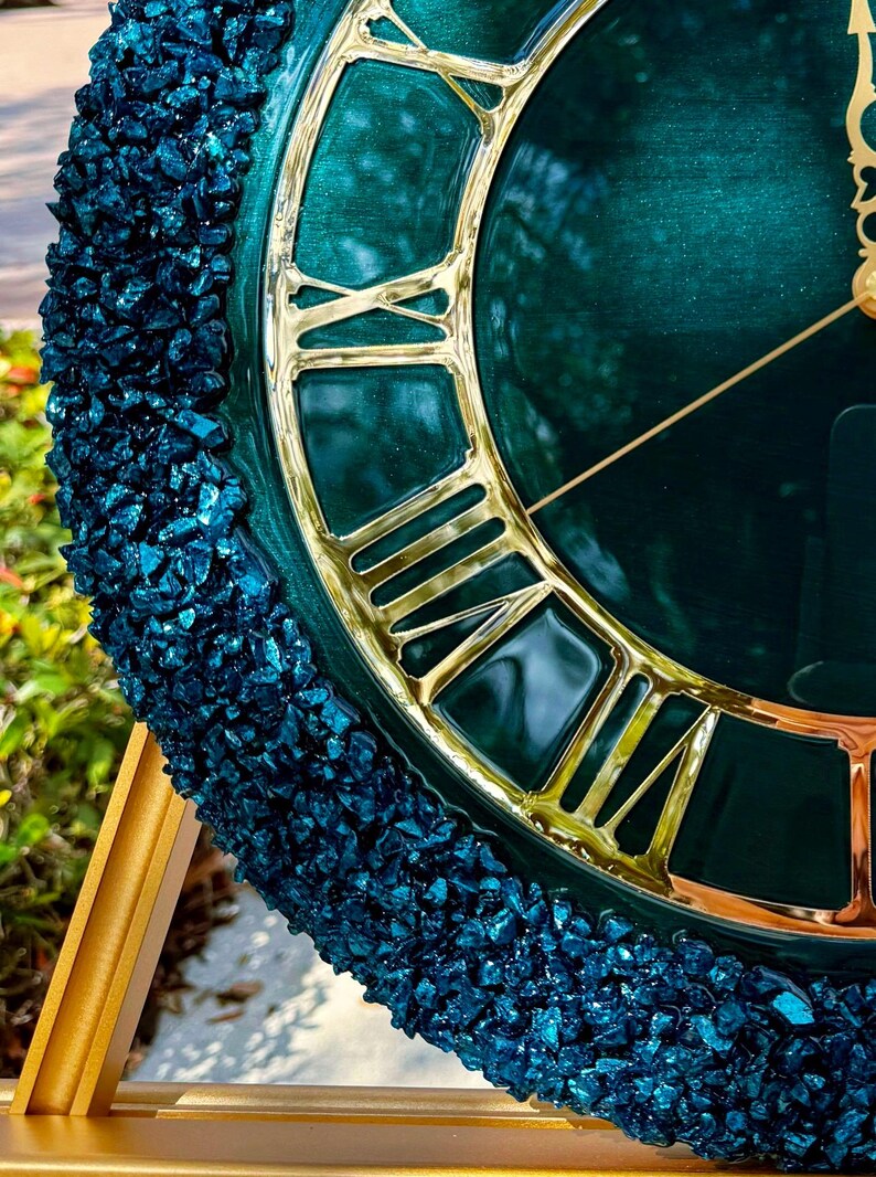 K&ouml;nnte beinhalten: Eine runde Wanduhr mit einem tief smaragdgr&uuml;nen Zifferblatt, polierten goldenen r&ouml;mischen Ziffern und passenden d&uuml;nnen goldenen Zeigern. Der &auml;u&szlig;ere Rand ist mit einer texturierten Schicht aus dunkelblauen Glassplittern besetzt.