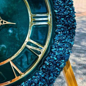 K&ouml;nnte beinhalten: Eine runde Wanduhr mit einem tief smaragdgr&uuml;nen, gl&auml;nzenden Zifferblatt, goldenen r&ouml;mischen Ziffern und verzierten Zeigern. Der &auml;u&szlig;ere Rand der Uhr ist mit einer dicken Umrandung aus strukturierten, dunkelblauen Glassplittern verziert.