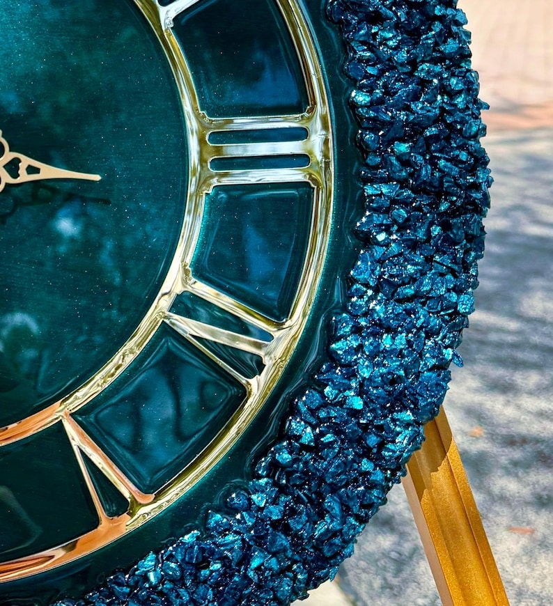 K&ouml;nnte beinhalten: Eine runde Wanduhr mit einem tief smaragdgr&uuml;nen, gl&auml;nzenden Zifferblatt, goldenen r&ouml;mischen Ziffern und verzierten Zeigern. Der &auml;u&szlig;ere Rand der Uhr ist mit einer dicken Umrandung aus strukturierten, dunkelblauen Glassplittern verziert.