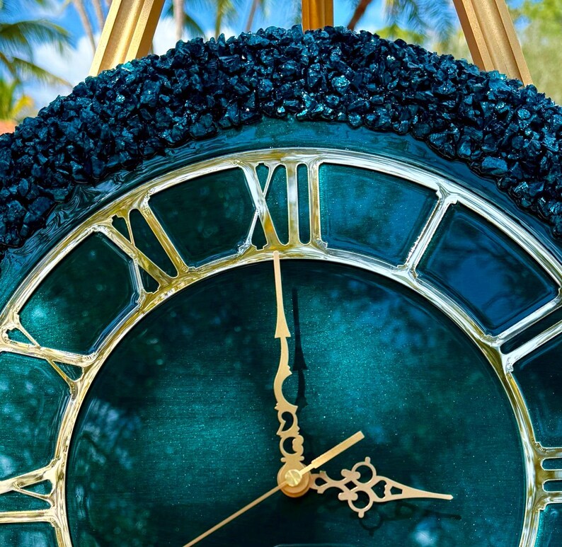 K&ouml;nnte beinhalten: Eine runde Wanduhr mit einem tief t&uuml;rkisfarbenen, schimmernden Zifferblatt und metallisch goldenen r&ouml;mischen Ziffern. Der &auml;u&szlig;ere Rand ist mit dunklen, strukturierten Steinen besetzt. Zwei verzierte, goldfarbene Metallzeiger zeigen die Zeit an.