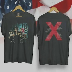 May include: Two black t-shirts displayed on wooden hangers. The front shirt features a band graphic with the letters "IN XS". The back shirt has a large red "X" with tour dates. The background includes an American flag.
