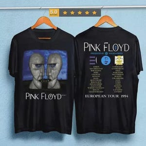 Puede incluir: Camisetas negras con gráficos de la banda Pink Floyd. La parte delantera muestra una imagen surrealista de dos caras, y la parte trasera enumera las fechas de la gira europea. Las camisetas están en perchas de madera.