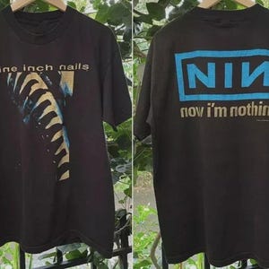 May include: Black Nine Inch Nails t-shirt with a graphic on the front and back. The front features the band's name and a stylized image. The back displays the NIN logo in blue and the phrase "now I'm nothing."