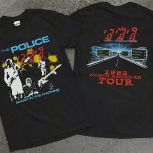 Könnte beinhalten: Zwei schwarze T-Shirts mit der Band The Police. Ein Shirt hat den Bandnamen in Blau und den Albumtitel "Ghost in the Machine". Das andere Shirt hat "1982 North American Tour" in Rot mit einer Straßen-Grafik.