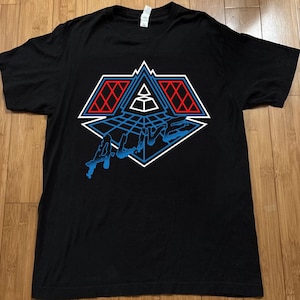 May include: A black short-sleeved t-shirt featuring a graphic print on the chest. The design includes a geometric pyramid shape with red grid patterns, white outlines, and the word ALIVE written in a stylised blue font across the bottom.