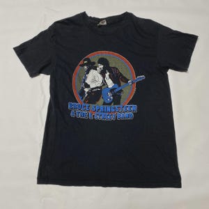 Op de afbeelding: Zwart T-shirt met een cirkelvormige afbeelding van twee muzikanten, waarvan één met een blauwe gitaar. De afbeelding staat in een rode, oranje en blauwe cirkel. De tekst "BRUCE SPRINGSTEEN & THE E STREET BAND" staat eronder.