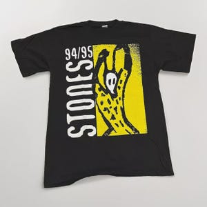 May include: Black t-shirt featuring a yellow and black graphic design with the text "STONES 94/95". The graphic is a stylized image of a figure with arms raised. The shirt is made of a soft material.