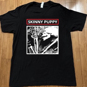 Puede incluir: Una camiseta negra de manga corta con un estampado gráfico blanco de una figura esquelética dentro de un marco cuadrado. Sobre el gráfico, el texto SKINNY PUPPY está impreso en letras mayúsculas blancas dentro de un borde rectangular rojo.