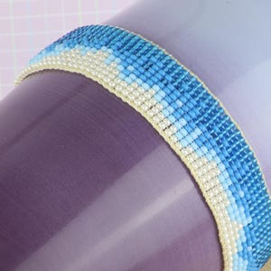 May include: A beaded bracelet with a gradient of blue and white beads. The bracelet is wrapped around a purple ceramic mug.