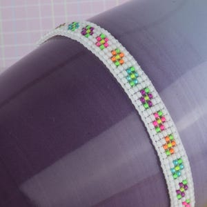 May include: A white beaded bracelet with colorful square beads in a repeating pattern. The bracelet is on a purple and white background.