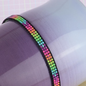 Peut inclure: Un bracelet en perles arc-en-ciel avec un fil noir. Le bracelet est sur une tasse à café ombrée de violet et de bleu.